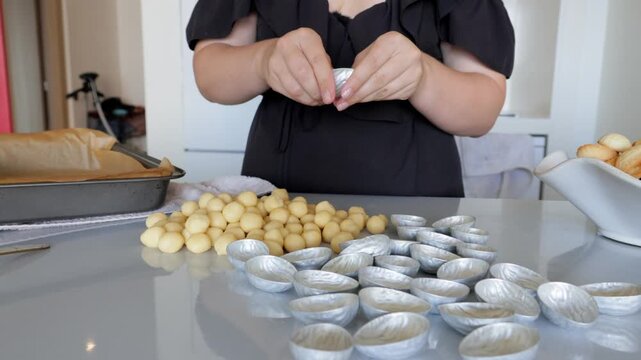 Hands forming tartlet centers on glossy counter, rows of dough balls and empty molds, bakery tray and mixing bowl visible, methodical pace and neat arrangement, indoor