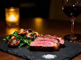 Sliced medium-rare steak served on a dark slate with saut��ed spinach and mushrooms, a pinch of salt, and a glass of red wine in a low-light setting.