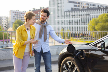 A couple stands near an electric vehicle while it is charging, with the man pointing at the charging port.