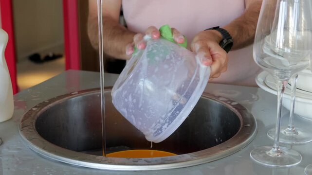 Caucasian man tipping plastic container to pour brown rinse water into round sink, steady hands and downward flow, kitchen counter with glasses at side, simple cleanup