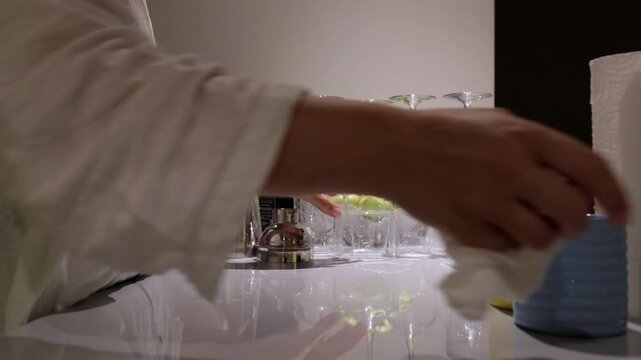 Person drying crystal glass beside fruit bowl, careful hand movements arranging stemware and cups on reflective countertop, ambient kitchen light and neat presentation