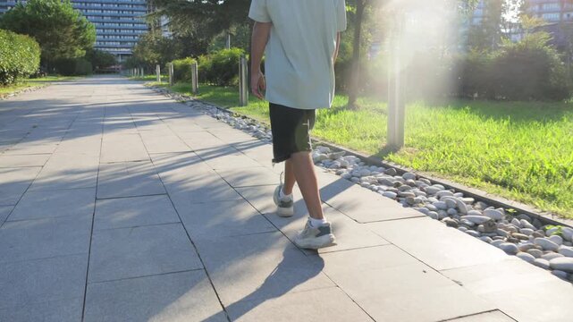 Youthful energetic stride, Dynamic young person moving swiftly amid scenic urban surroundings, Lively youth engaged in brisk walk beside landscaped areas under bright