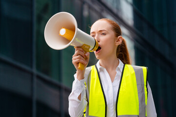 Young woman activist in yellow safety vest with megaphone loudspeaker leading protest action in...