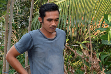 A relaxed man stands outdoors near coconut and banana leaves, gazing downward with focus and calmness, immersed in his peaceful rural environment and daily agricultural routine.
