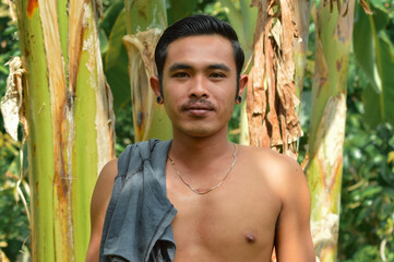 A young man poses confidently among banana trees, shirtless with a towel over his shoulder and necklace visible, bathed in warm natural light, reflecting rural pride and quiet resilience.