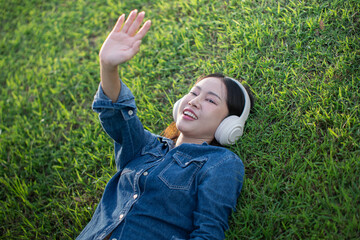 A beautiful Asian woman in a jeans shirt is sleeping lying on the grass, wearing headphones, and raising her hands to the sky happily.