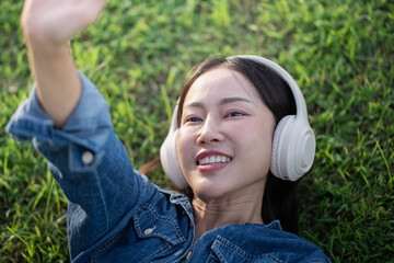 A beautiful Asian woman in a jeans shirt is sleeping lying on the grass, wearing headphones, and raising her hands to the sky happily.
