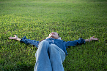 A beautiful Asian woman in a jeans shirt is sleeping lying on the grass, wearing headphones, happily.
