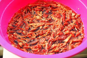 A top-down, close-up view of dried red chili peppers soaking and rehydrating in water inside a vibrant pink plastic bowl, with seeds floating on the surface.