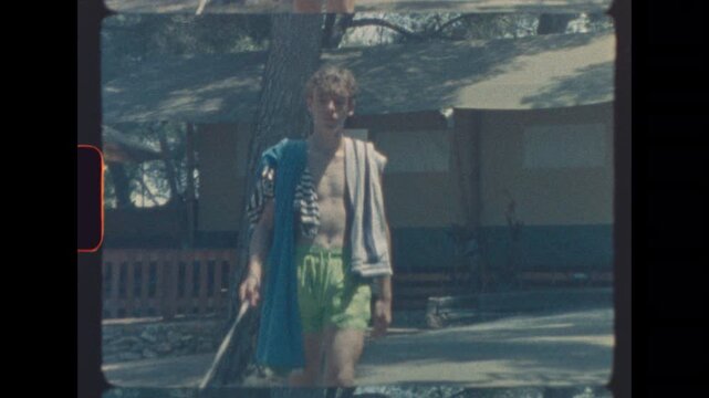 Shirtless teen in green swim shorts walking small dog on leash with towels over shoulder in sunny outdoor setting with retro film look