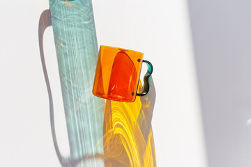 Orange Glass Mug with bright sunlight Shadows, transparent mug casting dynamic, vibrant lights reflections and shadows on light white surface, creative aesthetic top view, minimal close up