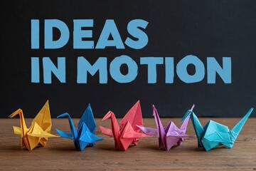 Colorful origami cranes on wood with "IDEAS IN MOTION" text on black background