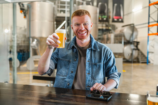 Smiling man with red hair holding a glass of fresh beer, enjoying a tasting session at a modern beer factory