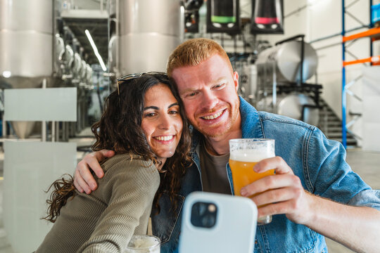 Happy young couple posing for a selfie with a glass of craft beer, enjoying a tour at a modern beer factory