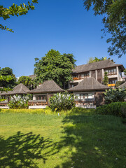 Serene Thai-style resort with lush tropical gardens and traditional wooden bungalows under a clear warm sky. Perfect for a tranquil getaway, featuring expansive lawns and vibrant foliage.