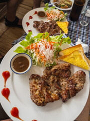 Delicious grilled pork steak served with a vibrant fresh mixed salad, crispy garlic bread, and a savory dipping sauce on a white plate. A perfect meal ready to be enjoyed.