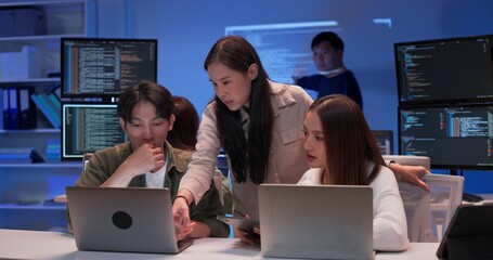 Focused group of young Asian developers working together on a coding project at night. Collaboration around laptops in a modern tech office with code screens and software development teamwork.