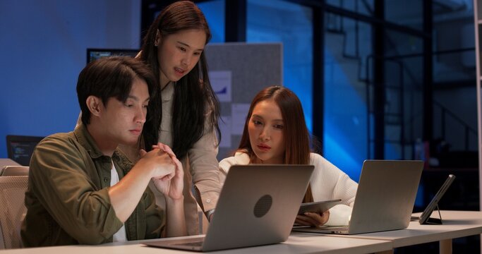 Focused group of young Asian developers working together on a coding project at night. Collaboration around laptops in a modern tech office with code screens and software development teamwork.