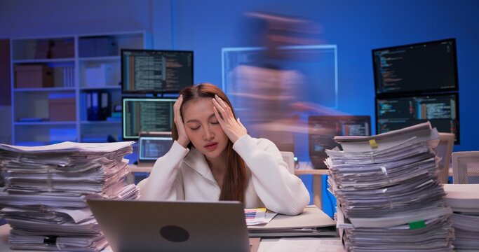 Asian businesswoman working overtime at modern office desk. Piles of paperwork grow rapidly as colleagues rush past. pressure and work overload. Young woman works late at night.