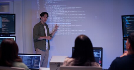Asian male developer presenting source code to colleagues in a dark tech office. Team listening to coding explanation during software training, AI session, or cybersecurity presentation.