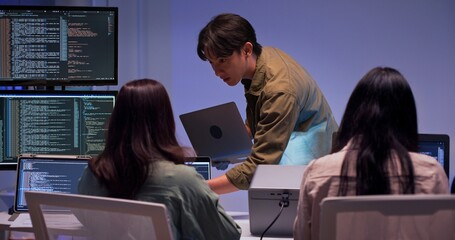 Group of young Asian developers collaborating during nighttime coding session. Team reviewing software issues and debugging source code in dark modern tech workspace. Innovation and teamwork in action