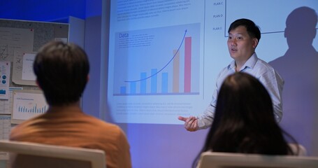 Confident Asian business professionals giving data-driven presentations to team in dark corporate office. Showing analytics charts, financial growth, and global strategy under projector light.