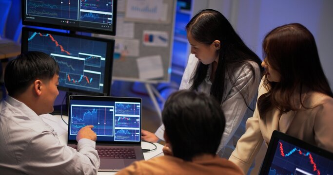 Asian business professionals working late night in modern office analyzing charts, discussing financial strategy, and reviewing market data across multiple monitors. Team collaboration and insight.
