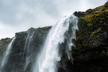 A powerful waterfall crashes down a rugged moss-covered cliff in Iceland under a moody sky. The force of the water and the dark, textured rock create a dramatic contrast, capturing the raw and untamed