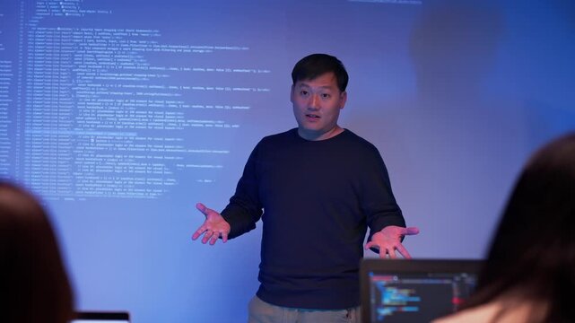 Asian male developer presenting source code to colleagues in a dark tech office. Team listening to coding explanation during software training, AI session, or cybersecurity presentation.