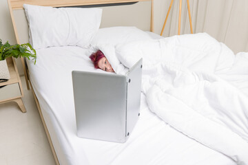 Asian young woman with purple hair, wrapped in blanket lying under white bed look at laptop computer in bedroom concept lazy weekend remote work relaxation comfort zone home lifestyle digital leisure.