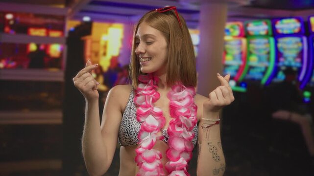Young blonde woman with bare arms forming a small heart shape with her fingers while smiling amid colorful slot machines in a casino; love affection happiness fun.