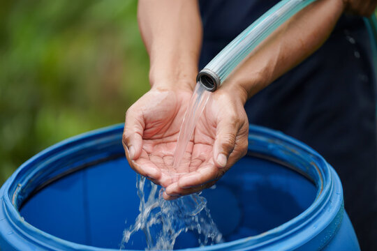Hands catching water from a hose filling a blue barrel, emphasizing water conservation and sustainable practices in a natural outdoor setting