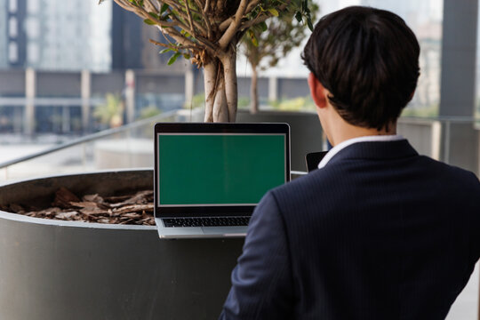 Young male professional working on a laptop with a green screen mockup outdoors at a modern office park. High quality photo