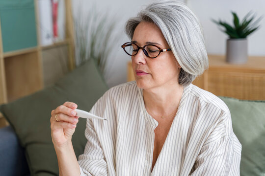 Woman holding thermometer with concerned expression at home