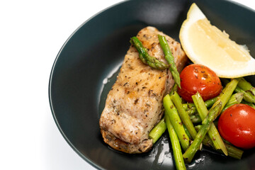 Red snapper steak, asparagus, tomatoes, and lemon on a black plate on a white background. Table top view.