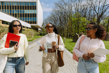 Two female students listen to a female student walking next to them while they hold notebooks and a cup and smile