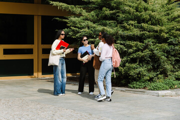 A group of four female students stand and talk while holding bags, folders, and notebooks