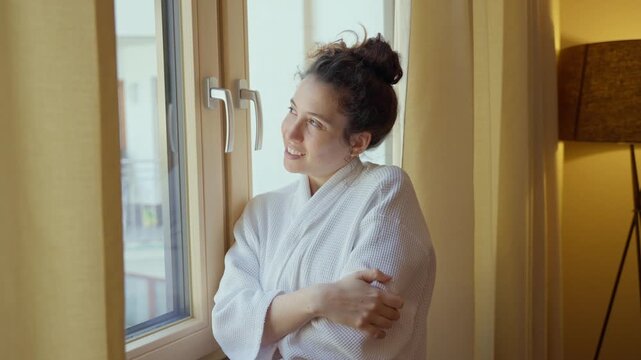 Young woman enjoys staying indoor in warm hotel wearing bathrobe looking outside. Female tourist in ski resort smiling inside cozy hotel suite with warm light standing by window during winter cold day
