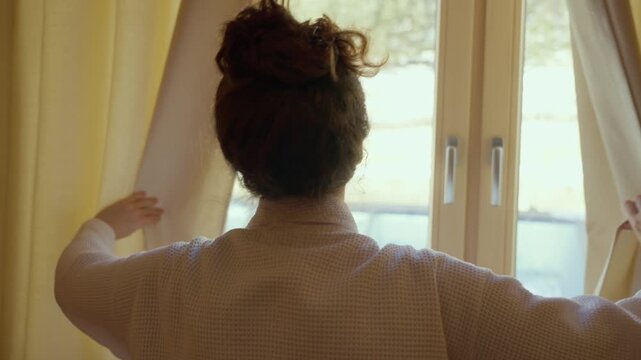 Female tourist hotel guest walks towards closed curtain or drape and draws open. Young woman enjoying spa resort or ski hotel suite open curtain in morning to reveal bright day outside with snow