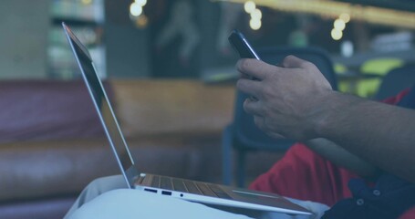 Holding smartphone, typing on laptop, man wearing dark t-shirt and red pants in lounge, copy space
