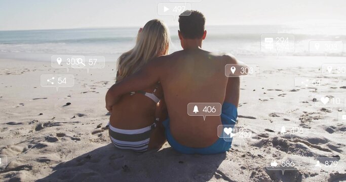 Sitting couple wearing swimwear embracing, watching sea on sandy beach, with social media icons - Powered by Adobe