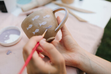 Painting ceramic mug with purple fish using brush and paint. Ceramic painting of mug with fish pattern using brush strokes. Artist paints delicate purple fish on a handmade clay mug.