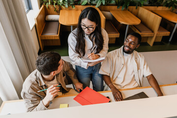 A female worker stands and writes in a notebook while listening to two male workers sitting next to...