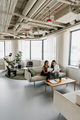 Two female employees are sitting on a couch and looking at documents that one of them is holding while a male employee next to them is looking at a laptop