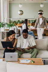 Obraz premium A female worker holds a laptop and sits on a couch next to a male worker who is talking while they look at the phone he is holding