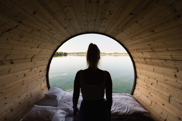 Silhouette of a woman relaxing on a bed in a wooden barrel cabin, looking at a tranquil lake view.