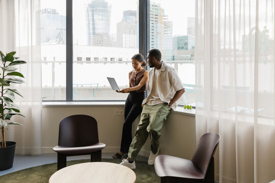 A male worker has his hands in his pockets and is talking to a female worker holding a laptop while they stand and lean on a windowsill