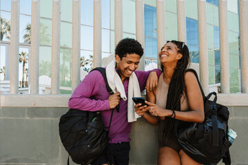 Male and female athletes laughing and leaning on a wall while he puts his hand on her shoulder and looks at the phone she is holding