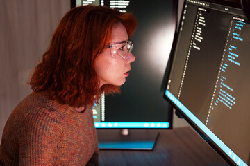 Developer wearing protective glasses focuses on code across dual wide screen monitors in a dim office, suggesting software engineering, debugging, and cybersecurity work.