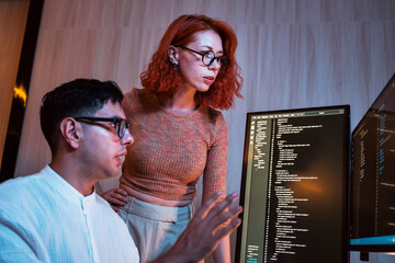 Two developers in a dim office review code on glowing monitors, collaborating on late-night programming. Scene shows software teamwork, startup energy, technology, planning, and coding in workspace.